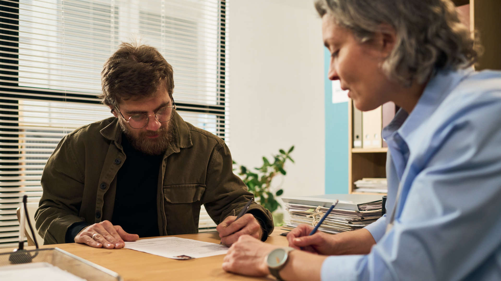 Ein Mann schreibt an einem Tisch auf einem Dokument, eine zweite Person sitzt ihm gegenüber und hält einen Stift.