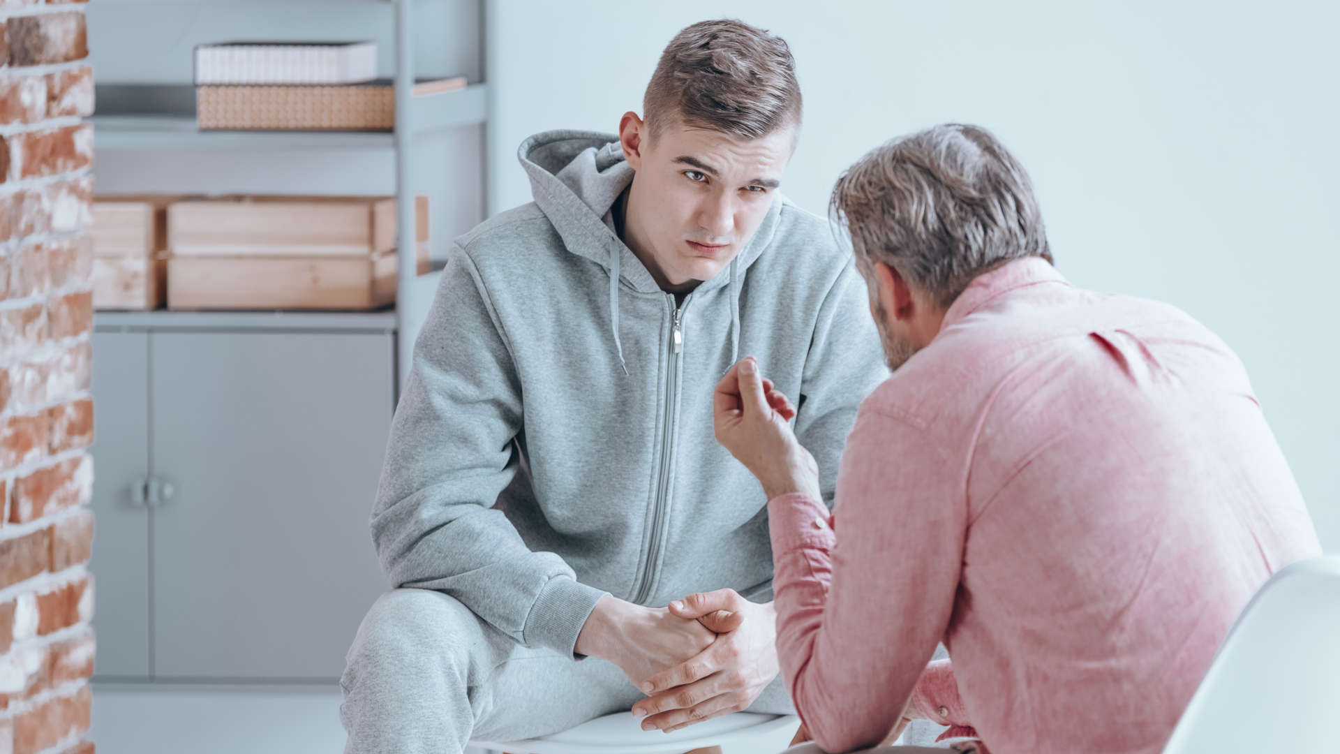 Ein junger Mann sitzt mit verschränkten Händen auf einem Stuhl, ihm gegenüber sitzt eine weitere Person im Gespräch.