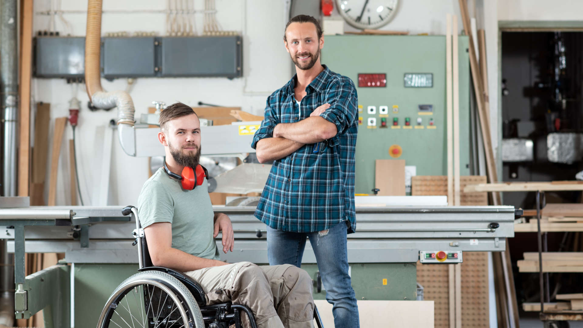 Ein Mann im Rollstuhl und ein weiterer Mann stehen gemeinsam in einer Werkhalle vor einer großen Arbeitsmaschine.