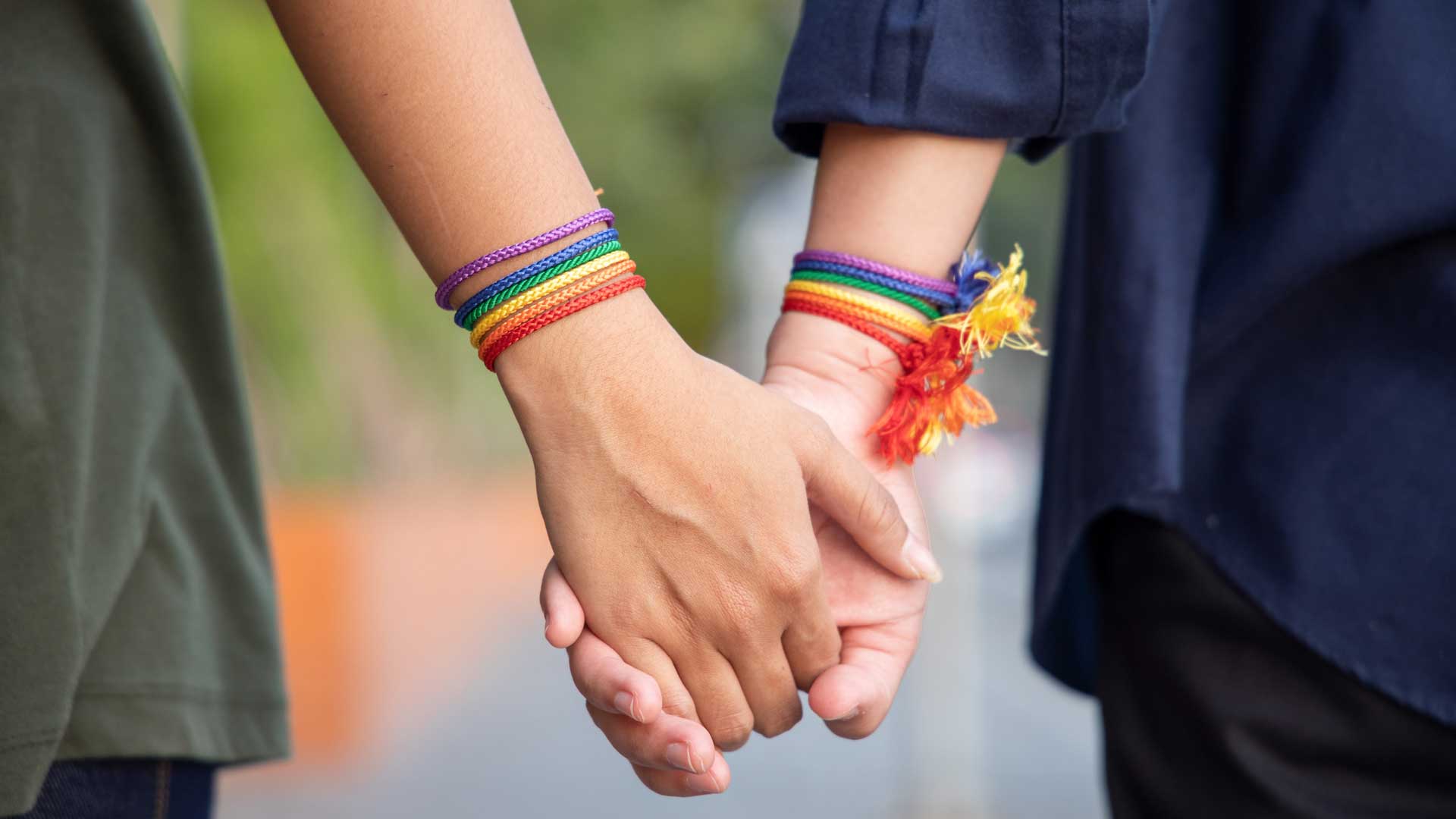 Zwei Personen halten sich an den Händen, beide tragen Armbänder in Regenbogenfarben.