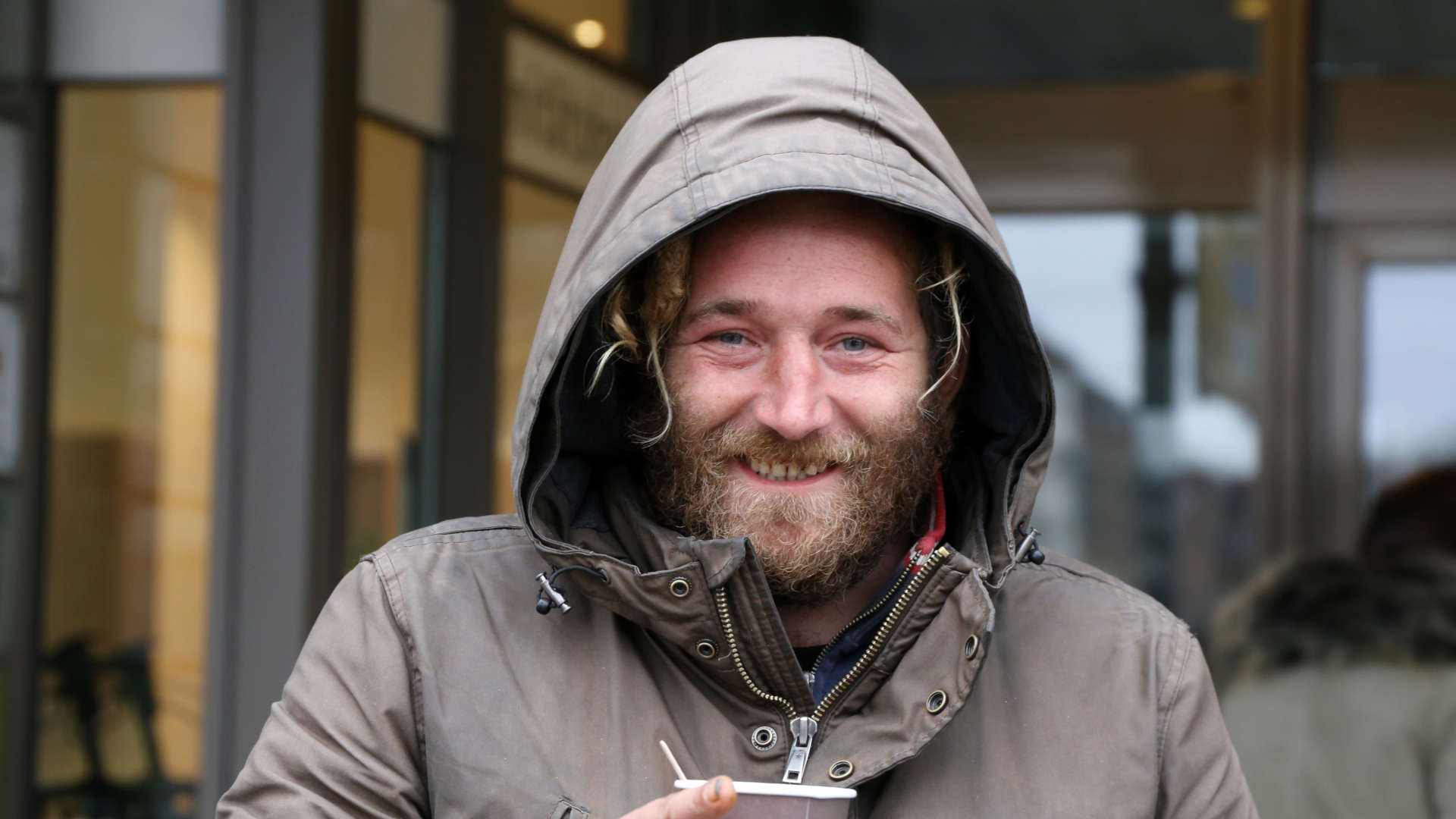 Porträt eines Mannes in wetterfester Jacke mit Kapuze, der vor einem Hauseingang einen Becher hält