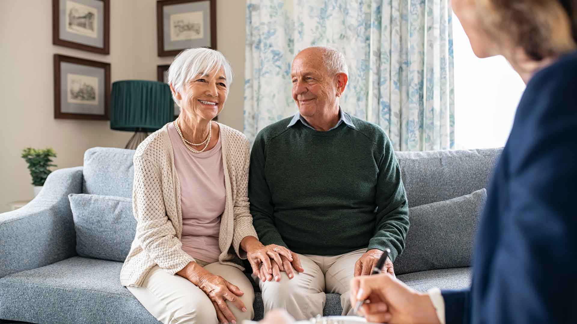 Ein älteres Paar sitzt lächelnd auf einem Sofa und blickt zu einer weiteren Person, die im Vordergrund angeschnittenzu sehen ist.